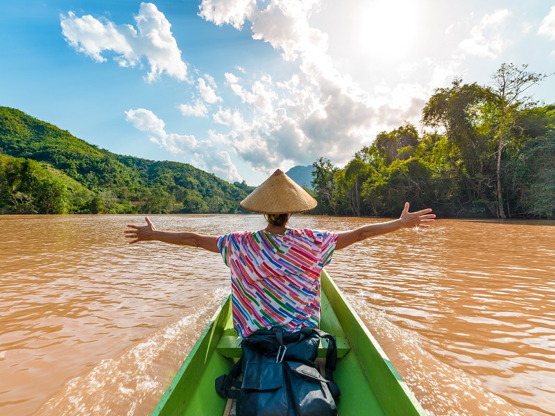 Woman with traditional hat cruising on the brown water of the Nam Ou river in Laos, amazing landscape mountain jungle famous travel destination in South East Asia rear view